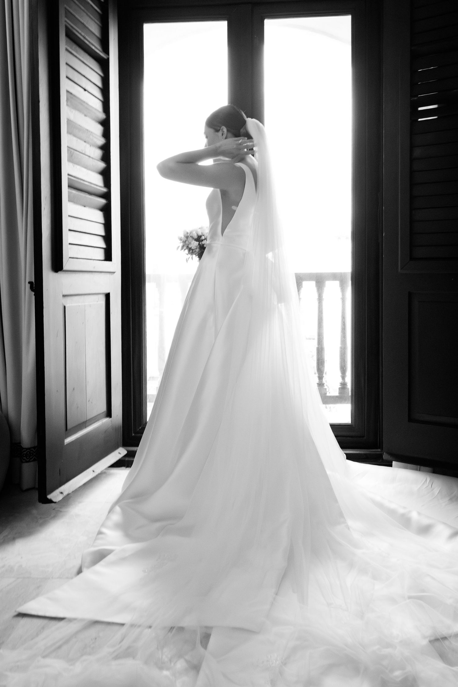 A bride in a flowing white wedding gown stands silhouetted in a doorway holding a bouquet, creating a dramatic backlit effect.