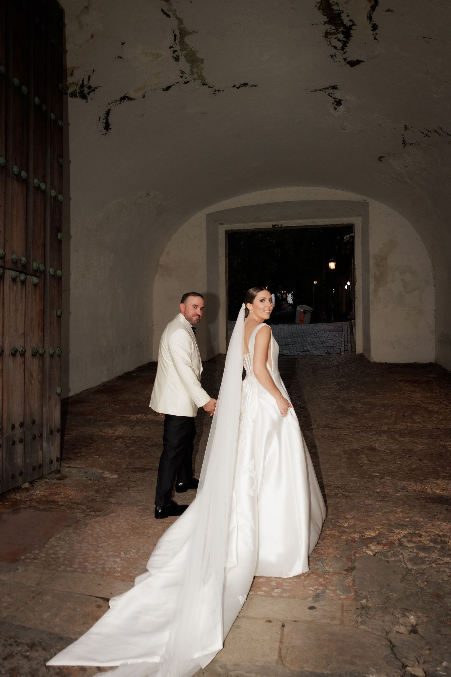 A couple in formal wedding attire poses in a rustic stone archway with dramatic lighting.