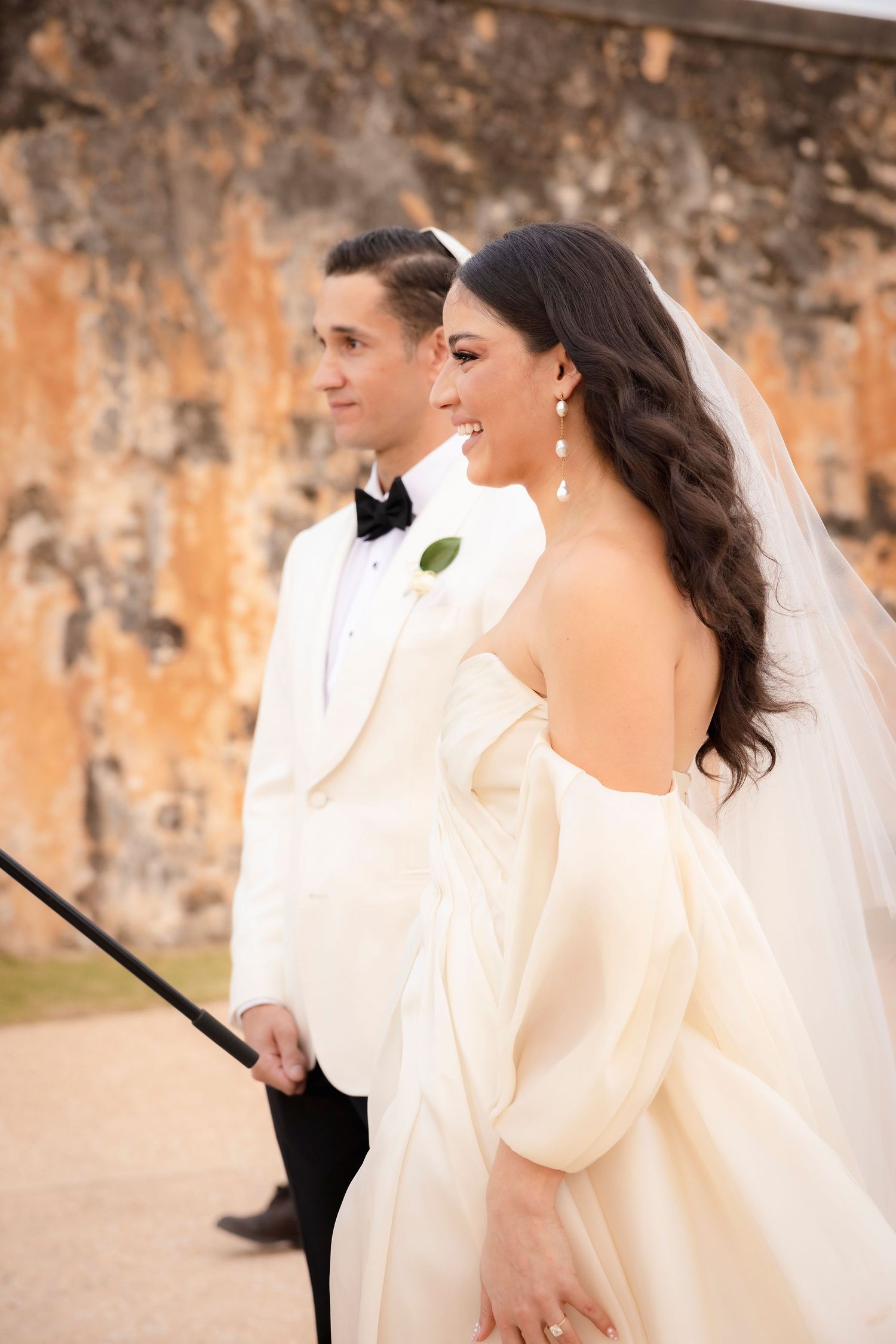 Elegant couple in white formalwear walking against textured stone wall backdrop.