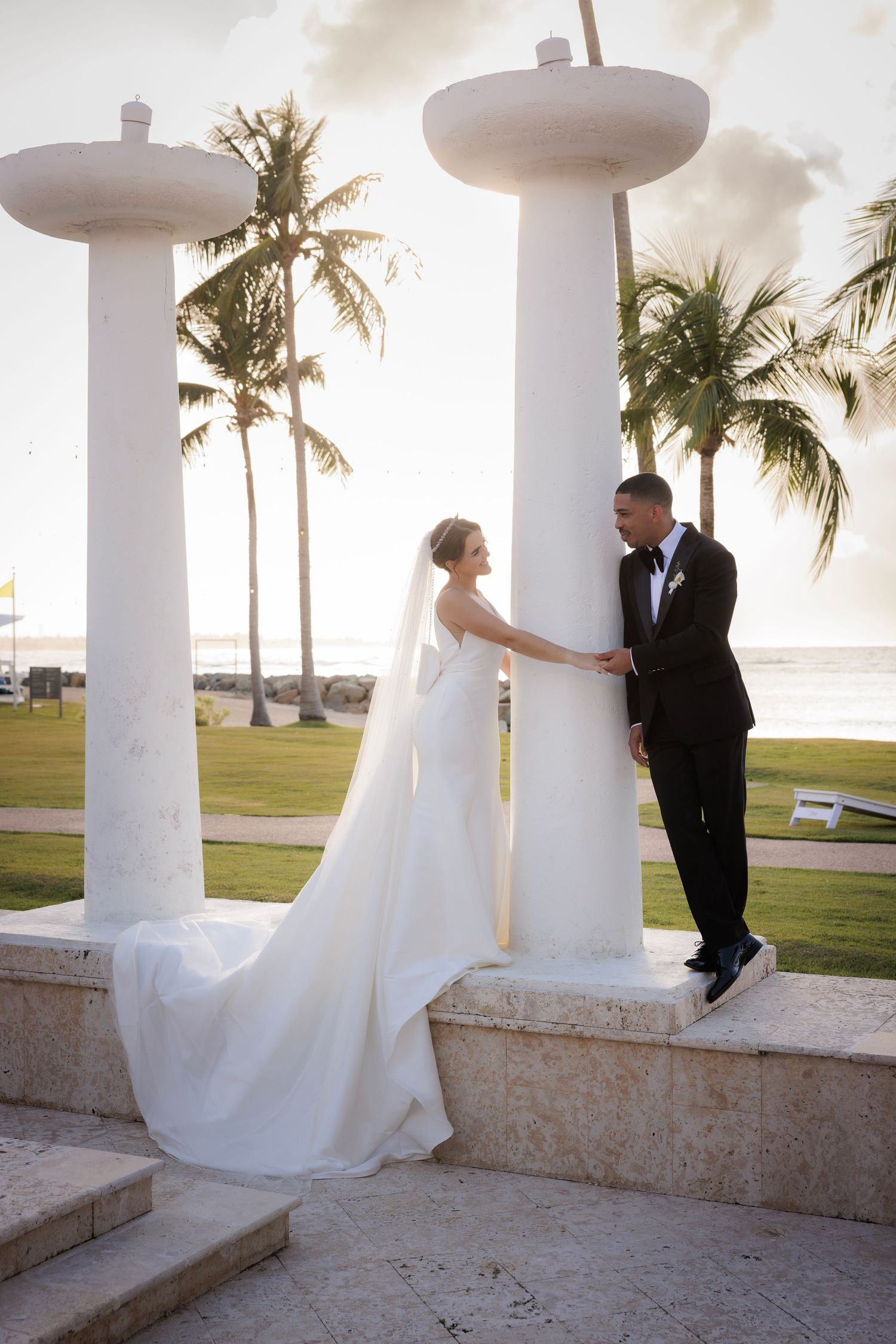 Romantic wedding couple poses together between white columns on a tropical beachfront at sunset.