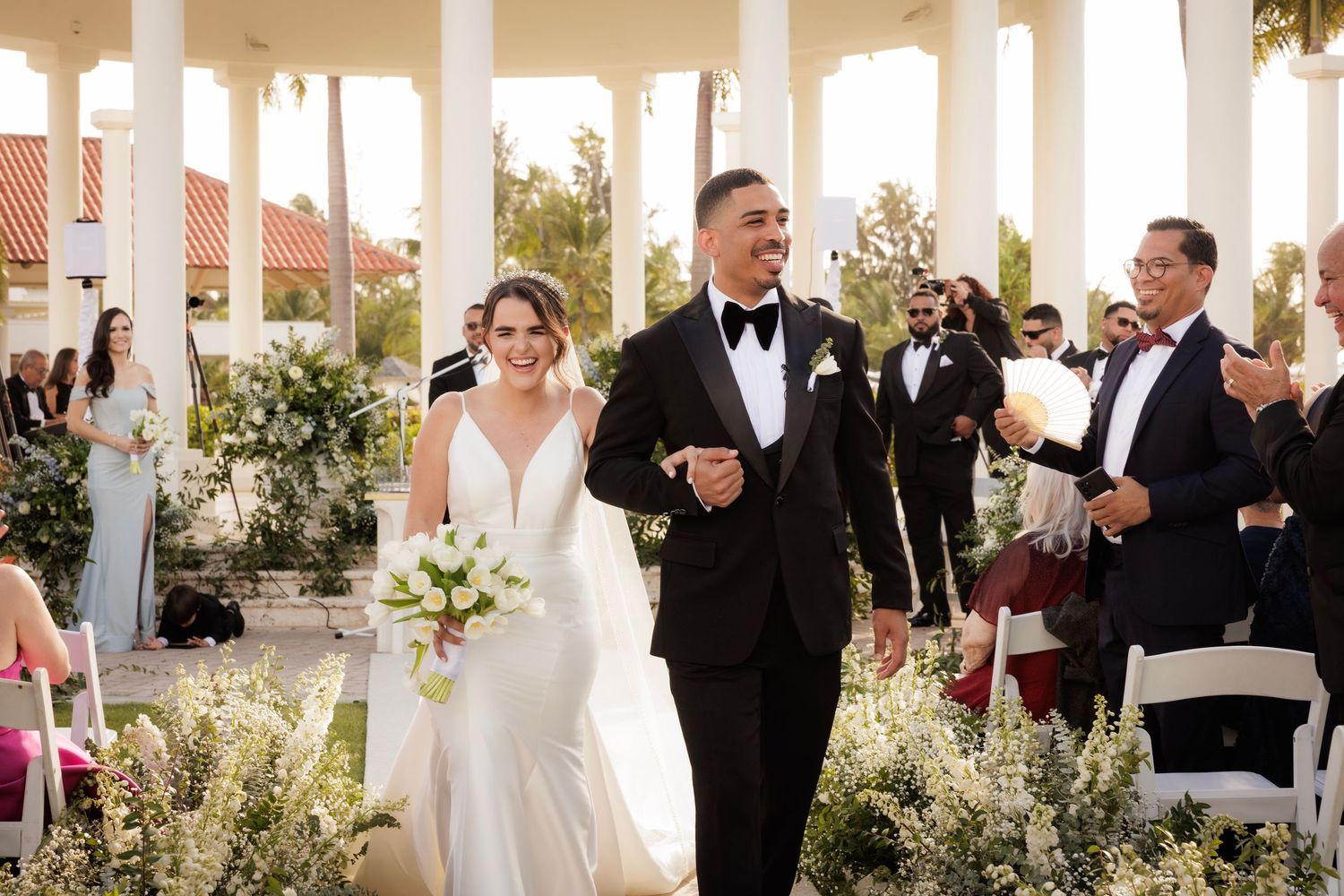 Outdoor wedding celebration with guests mingling on an elegant columned terrace decorated with floral arrangements.