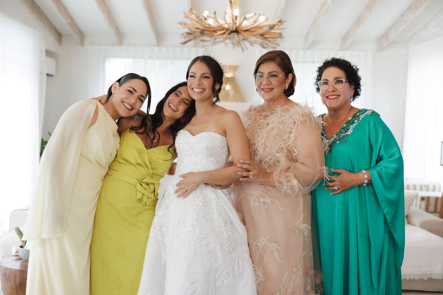 A bridal party in pastel yellow and turquoise dresses share joyful moments in a bright white room.