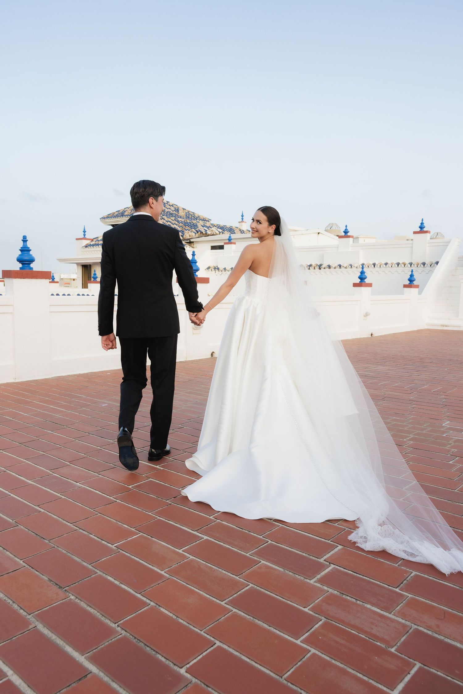 A newlywed couple walks hand in hand on a terracotta tiled rooftop against a blue sky.