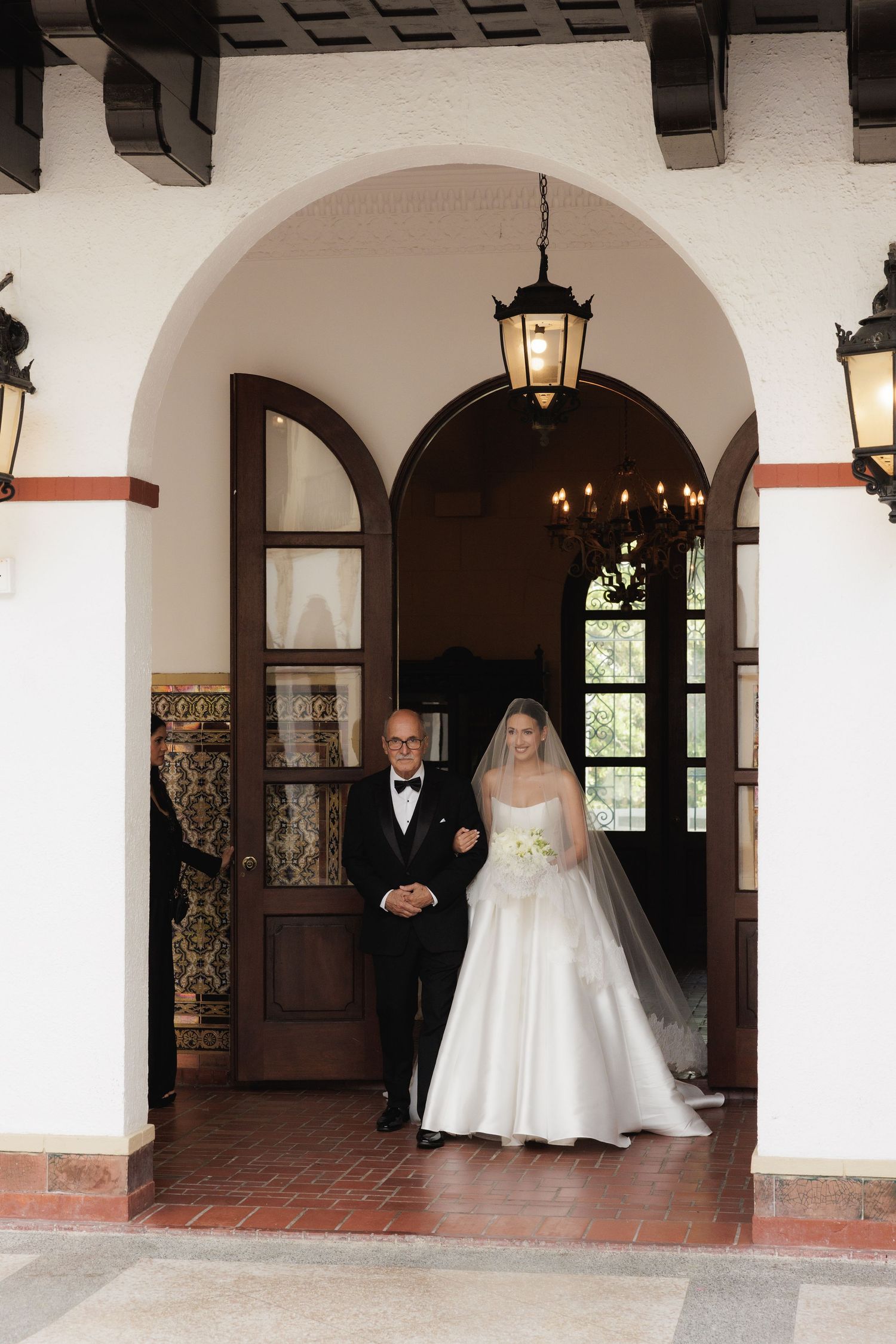 A bride in a white wedding gown walks through an elegant Spanish-style archway corridor with hanging lanterns.