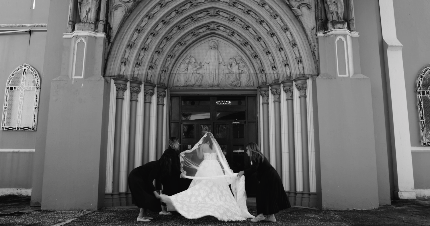 Black and white photo of a bride in a white gown with veil being arranged by attendants at ornate church doors.
