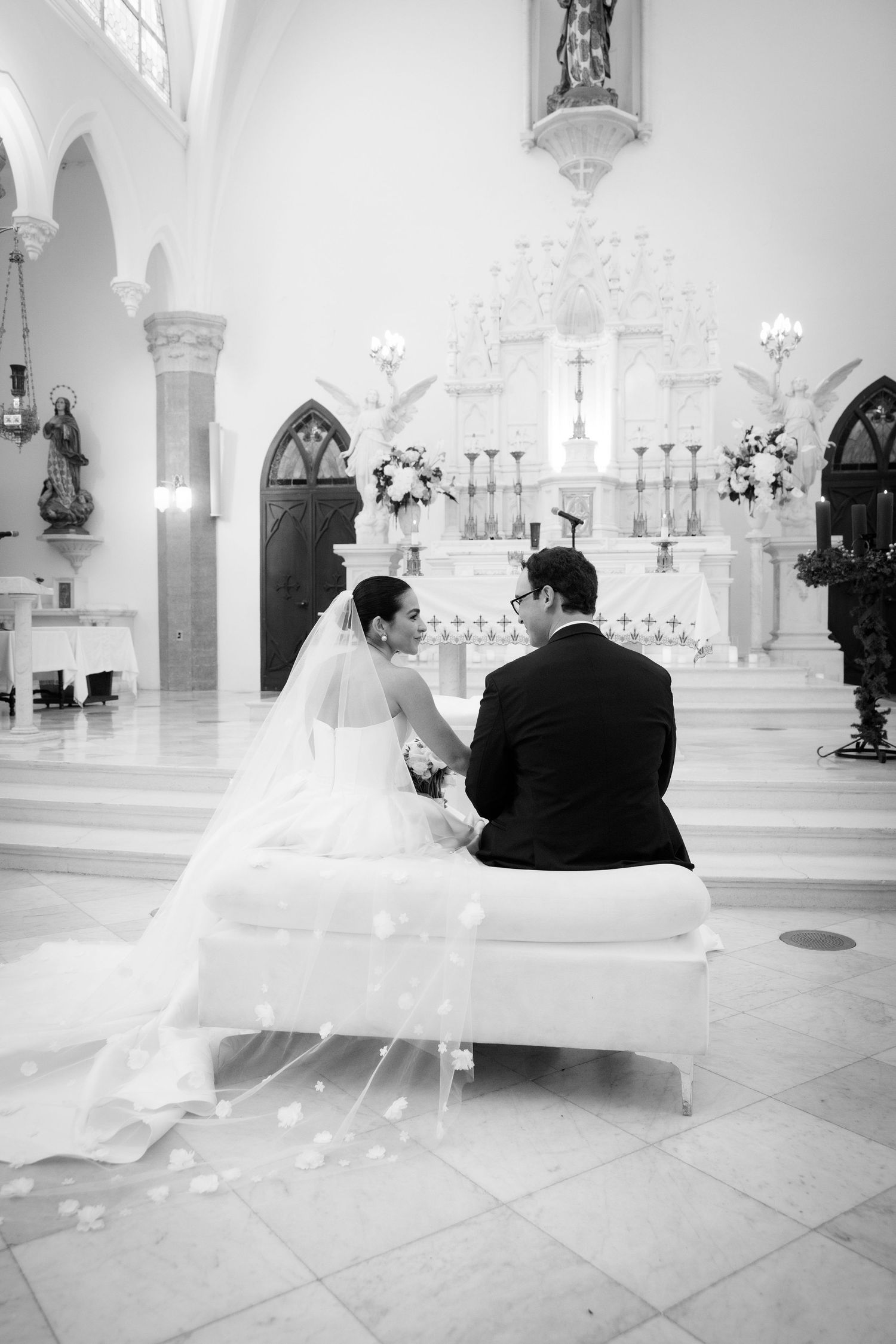 A couple sits together at the altar of an elegant white church during their wedding ceremony.