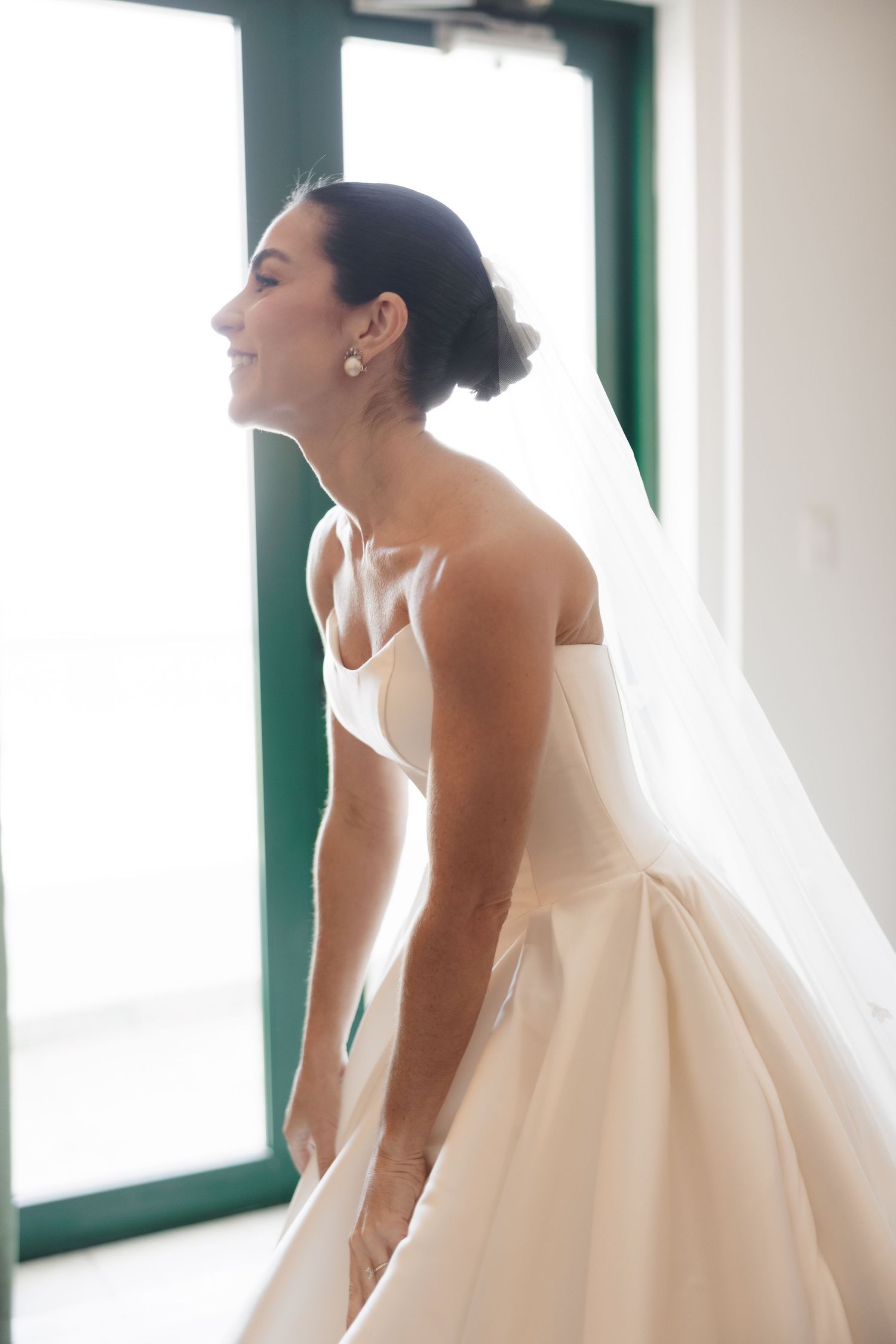 A bride in a white strapless wedding gown laughs joyfully near windows with green trim.