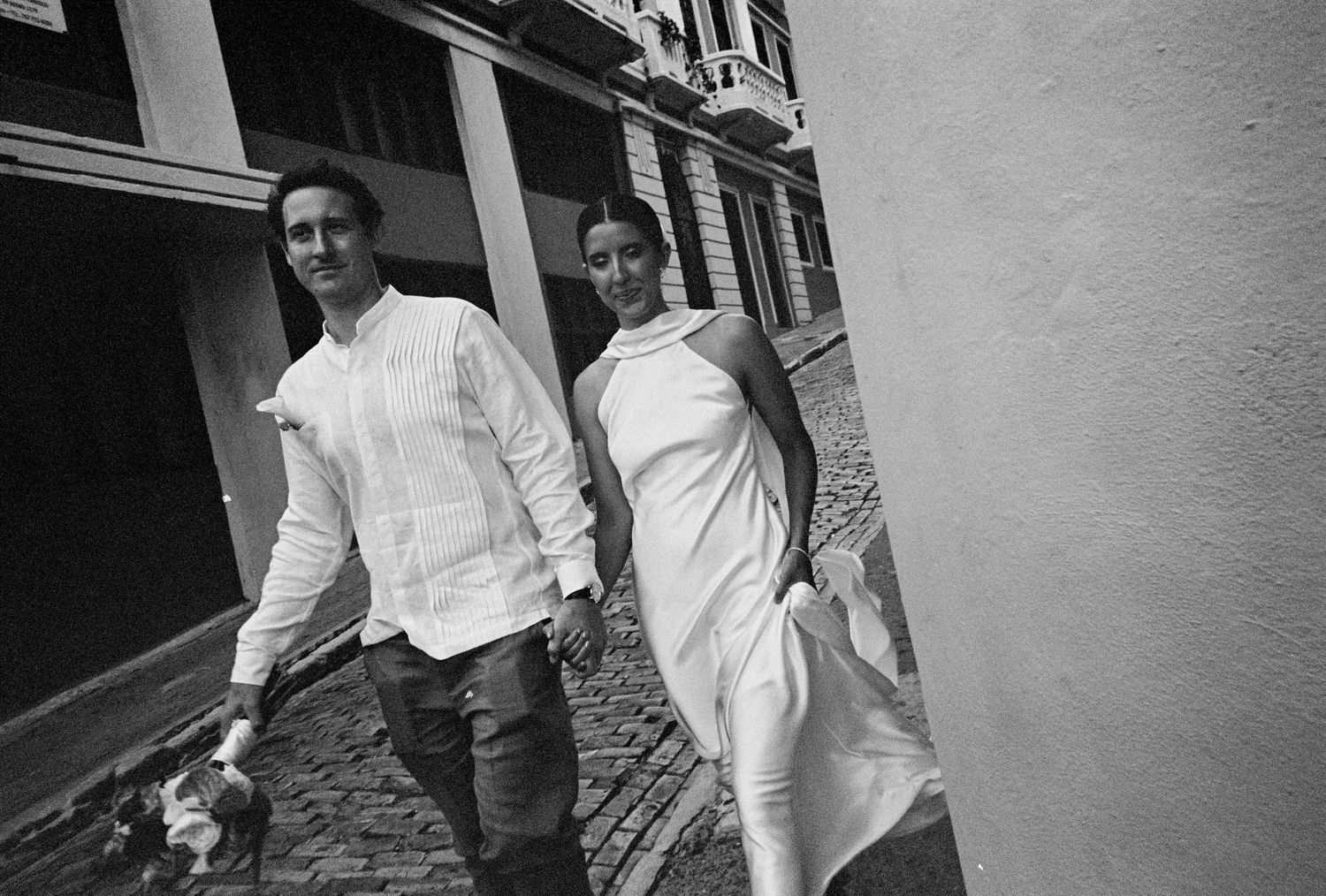 Black and white photo of a man and woman walking on cobblestone street, man holding a small dog.