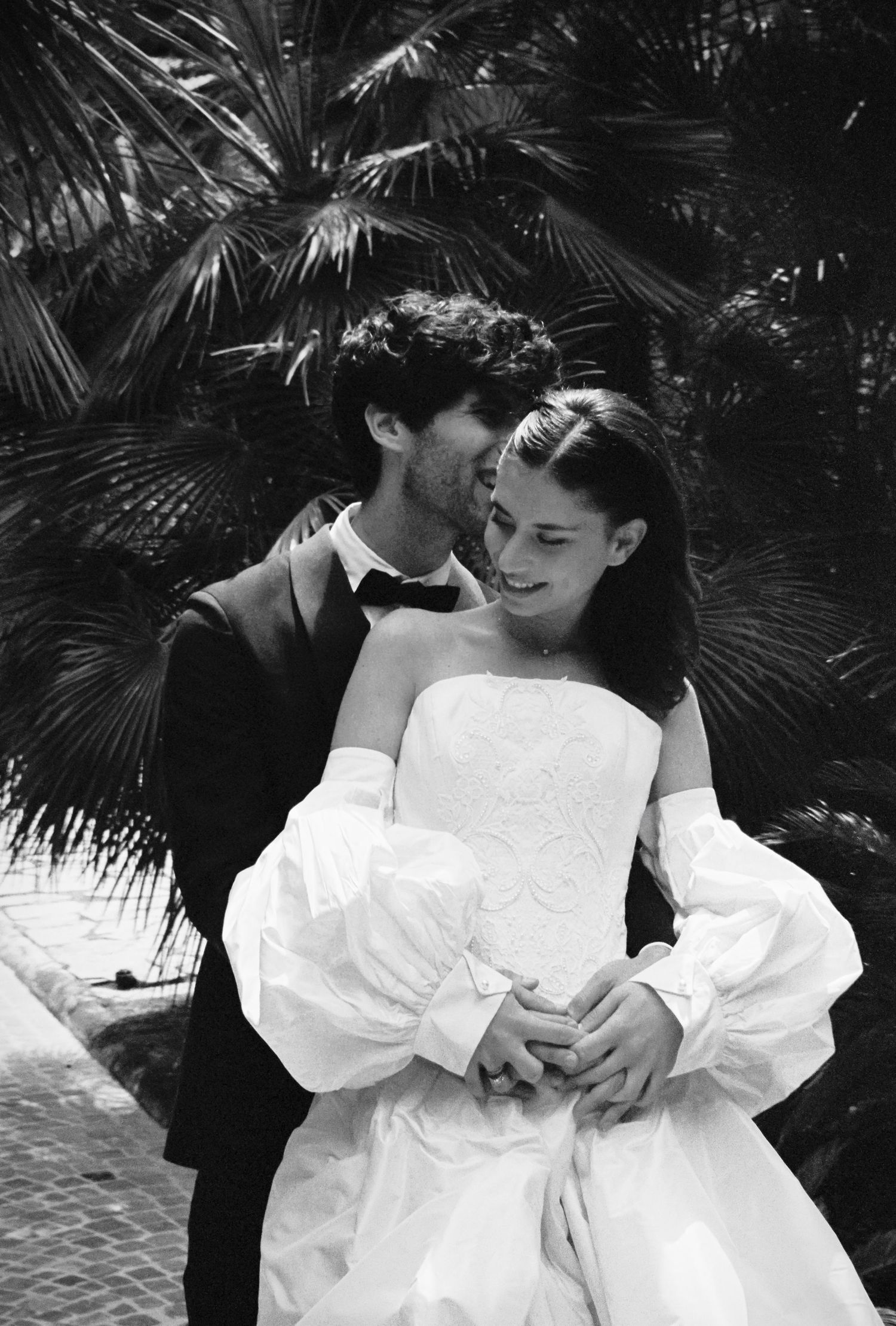 Black and white wedding photo of smiling bride in white gown with groom in suit embracing near palm trees.
