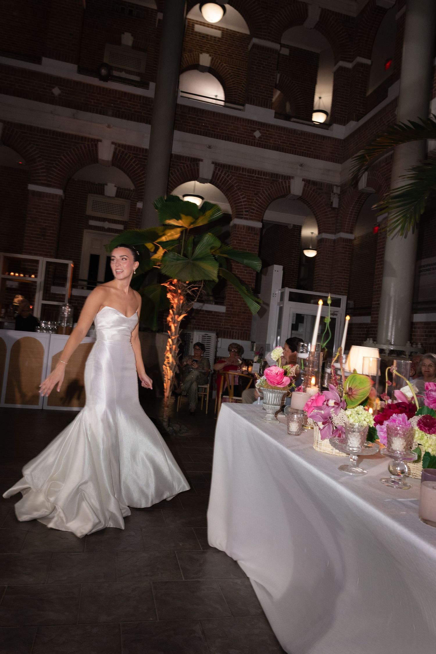 Bride in white mermaid gown walking near floral centerpieces at elegant wedding reception in brick venue.