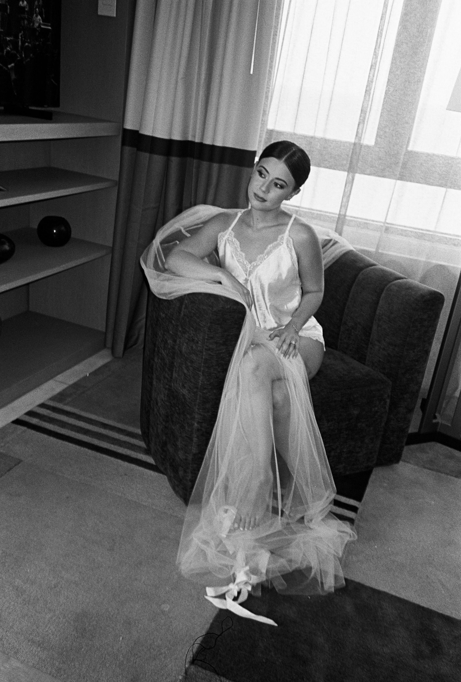 Black and white photo of a bride in a flowing white negligee and robe sitting elegantly in an armchair.