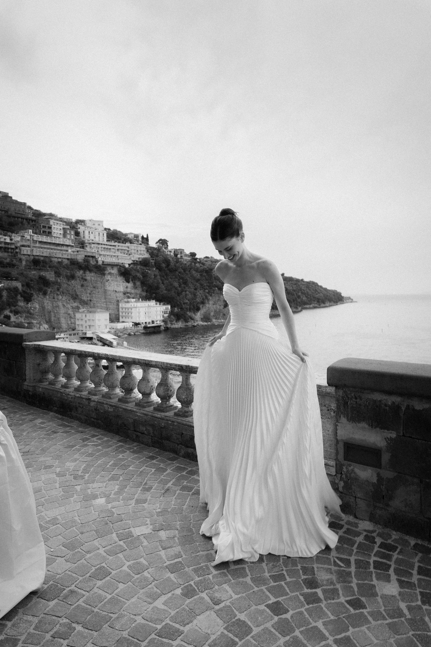 Bride in white pleated gown on coastal terrace with sea views, black and white wedding photo.
