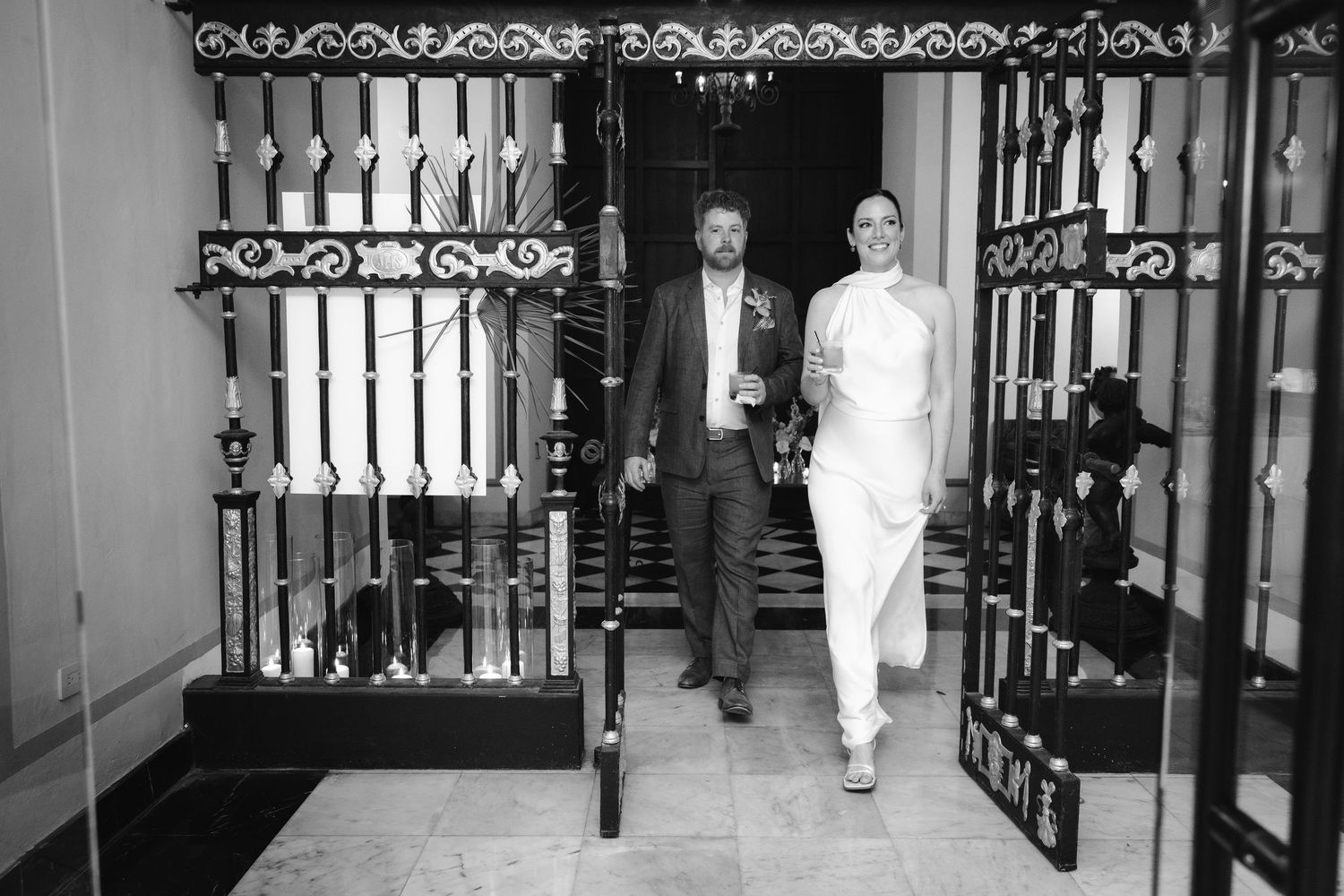 Black and white security camera footage shows a person walking through a dark hallway with ornate iron gates along the walls.