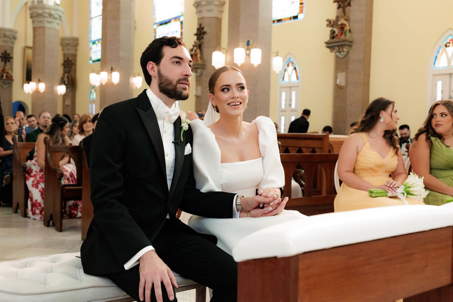 A wedding couple sits in church pews surrounded by guests and ornate architecture during a ceremony.