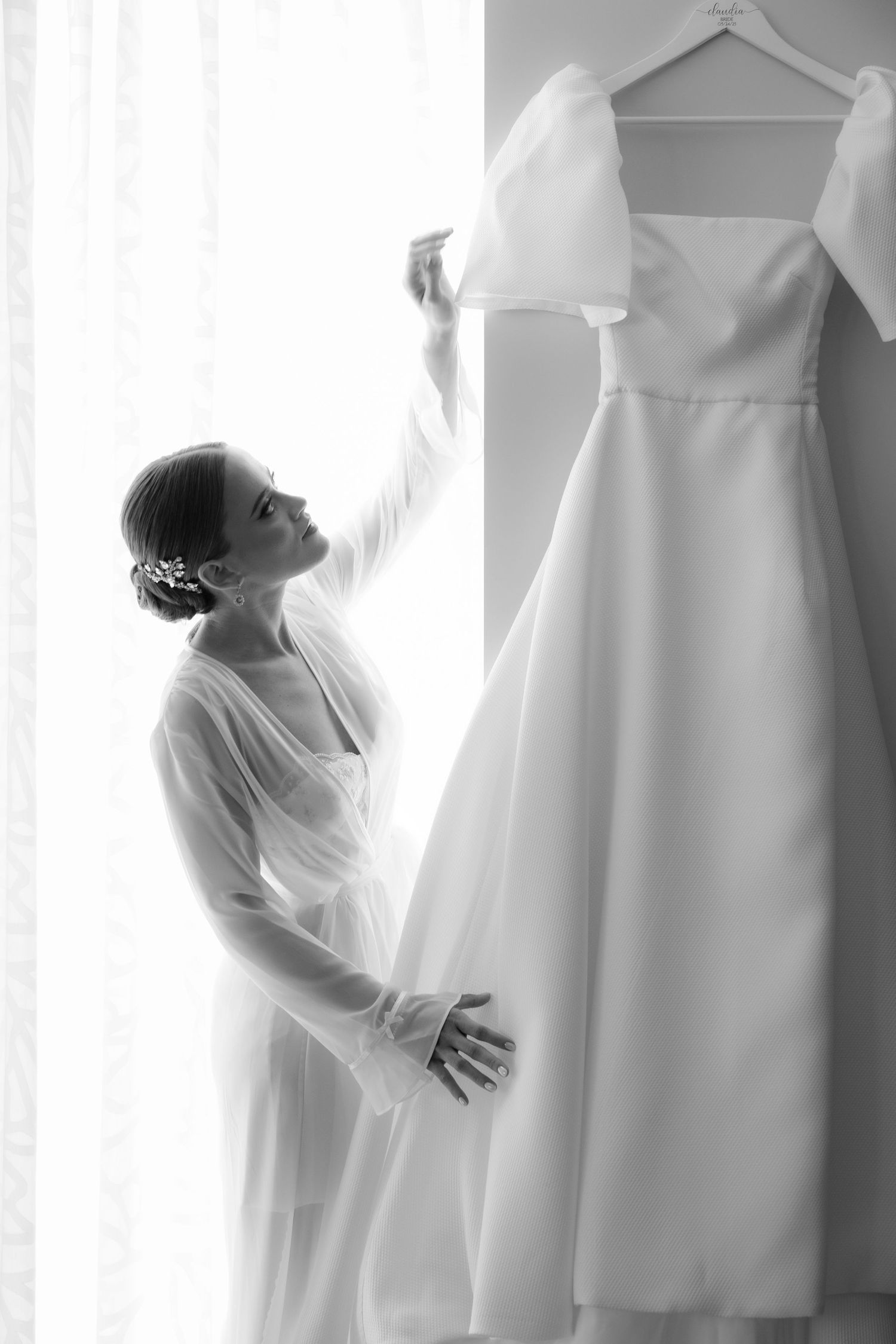 Black and white photo showing a wedding dress hanging while being admired beside a window in an intimate morning moment.