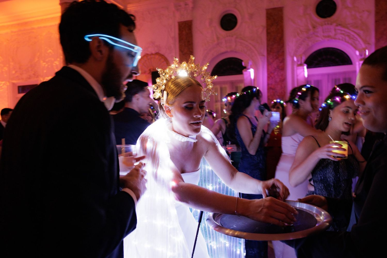 Party guests in festive attire enjoy the dance floor under pink and orange lighting at an indoor celebration.