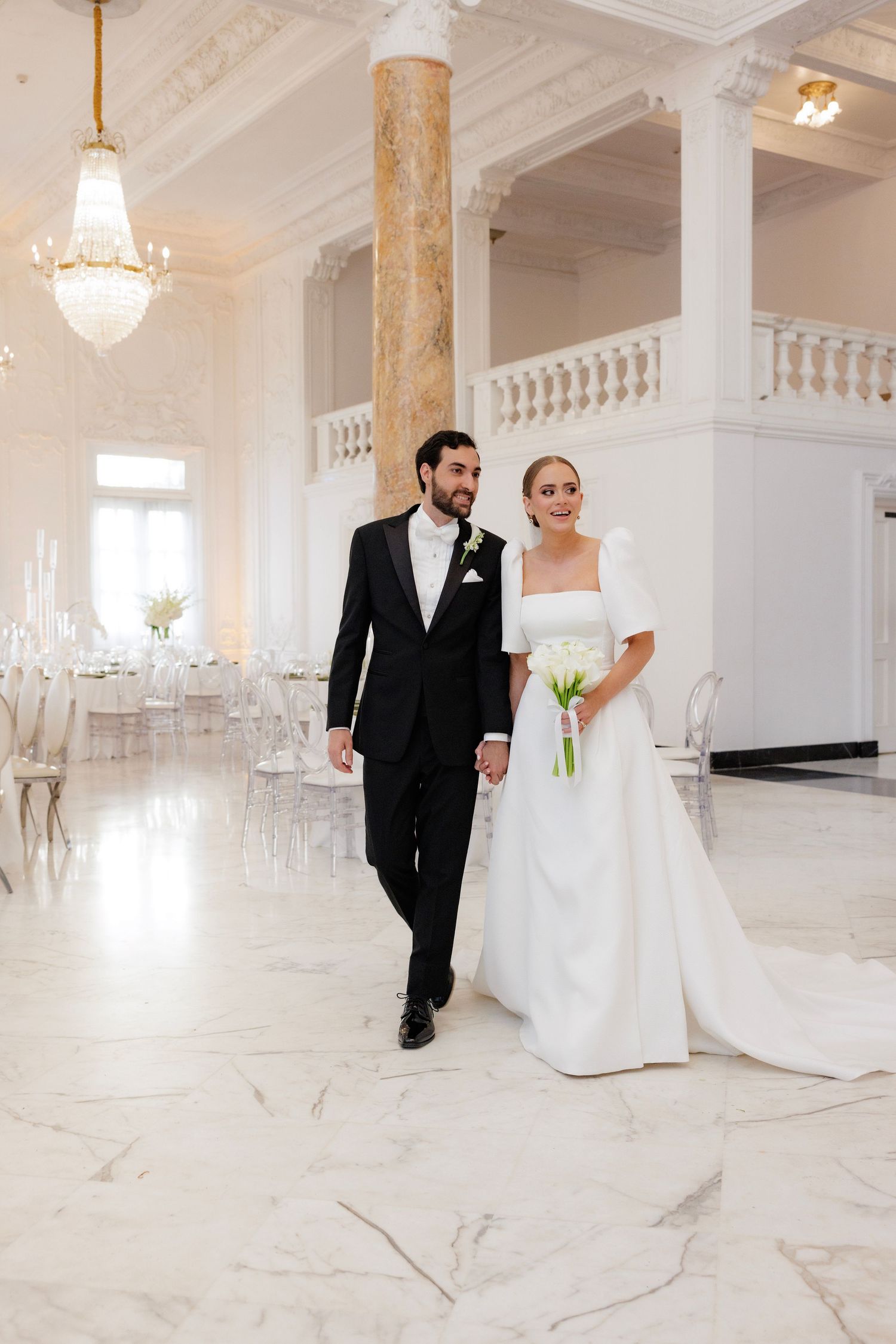 A bride and groom walk together in an elegant white marble ballroom with crystal chandeliers and classic columns.