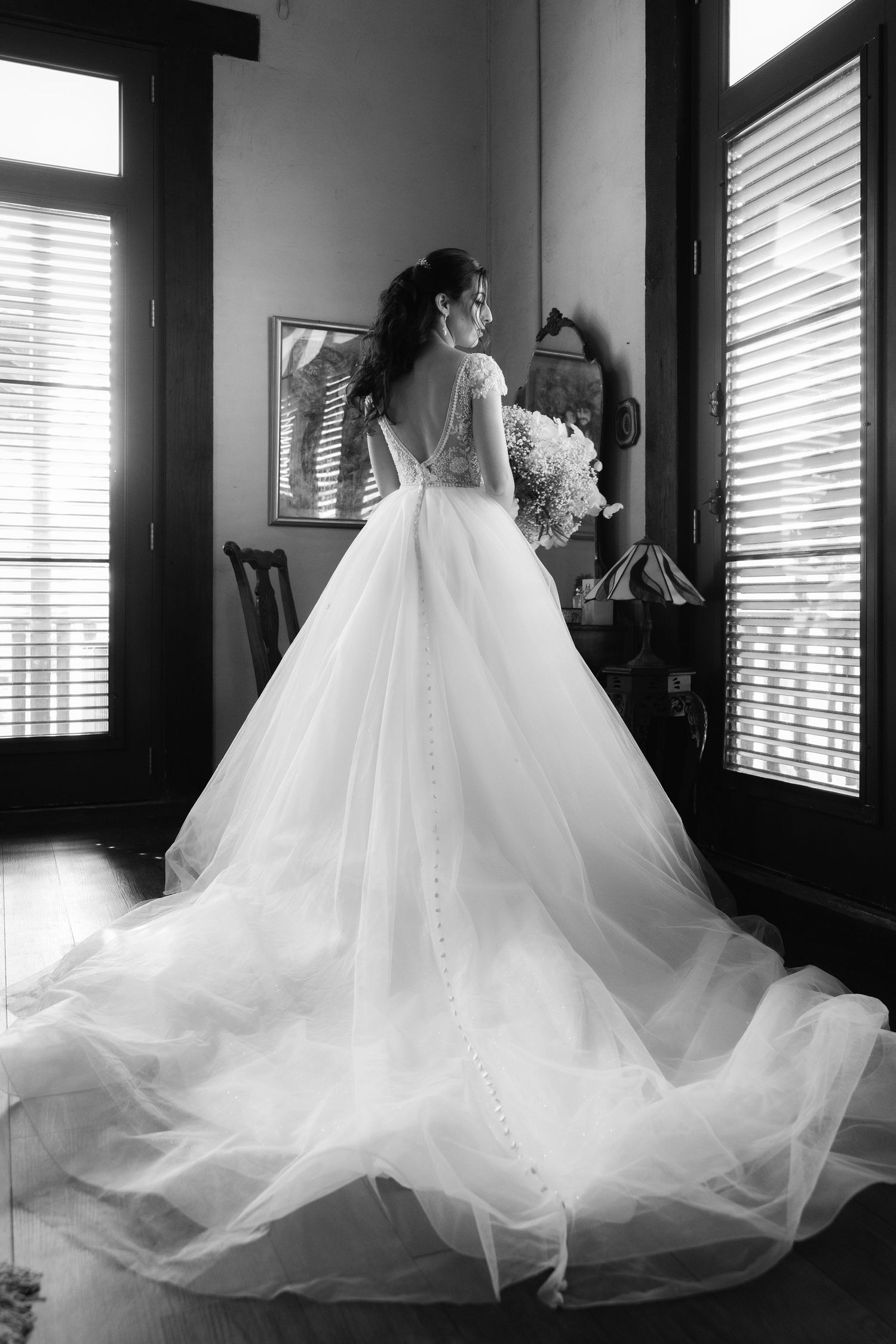 Black and white photo of a wedding gown with dramatic flowing tulle train by window light.