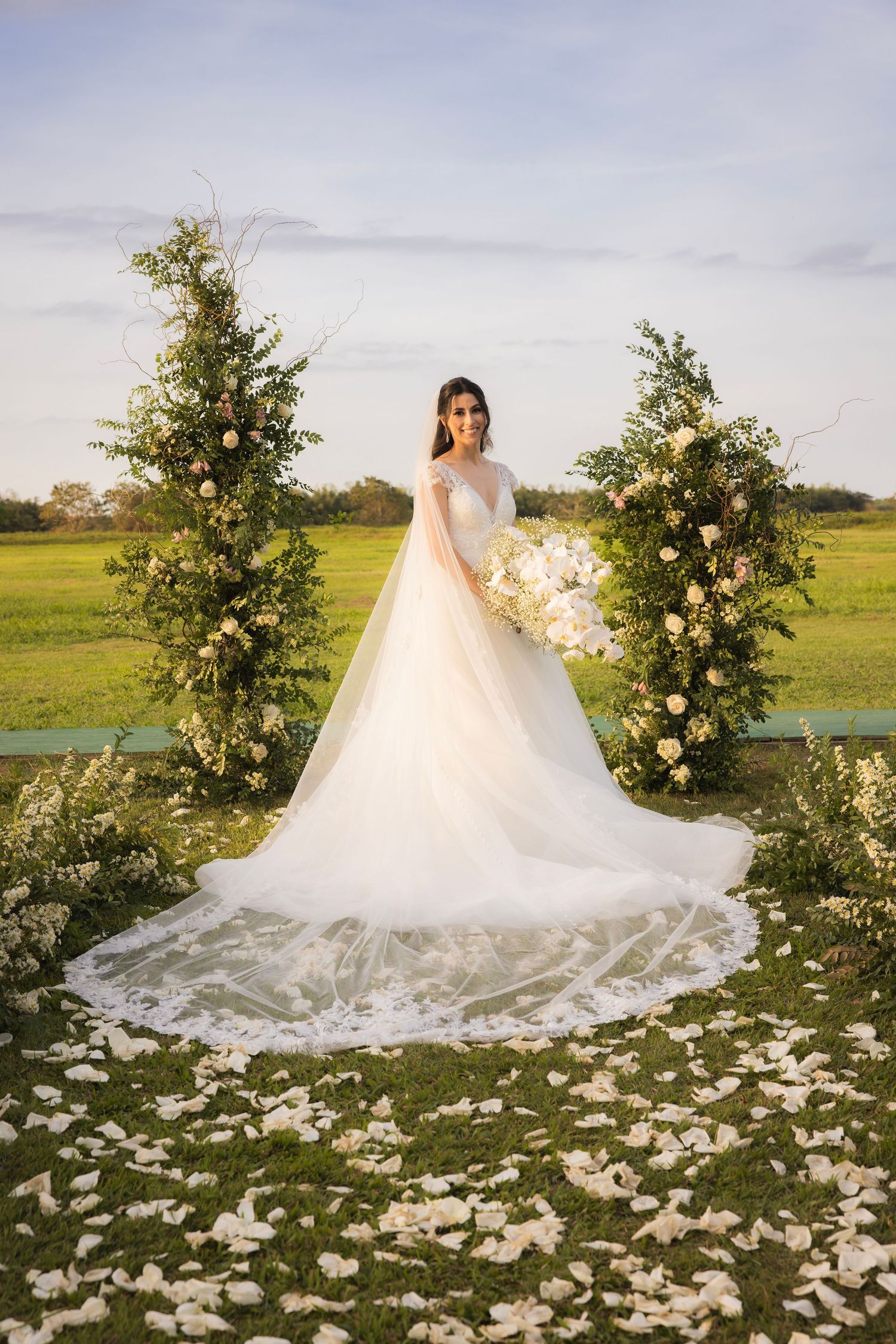 A flowing white wedding gown with a long train spreads across a grassy field decorated with flower petals and floral arrangements.