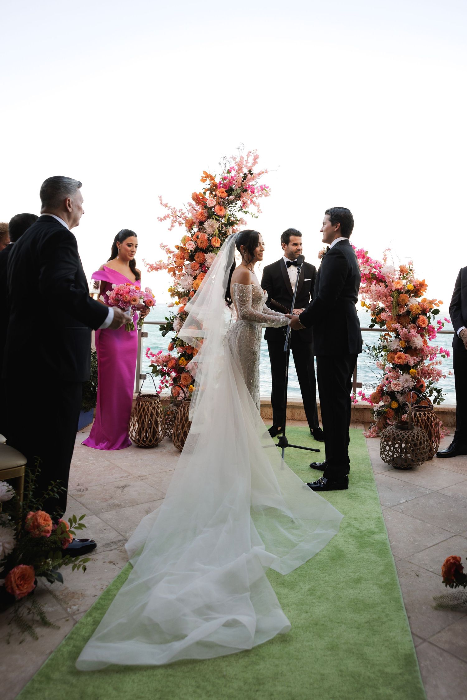 Wedding ceremony in progress with couple and guests gathered on green carpet under floral arch with orange blooms.