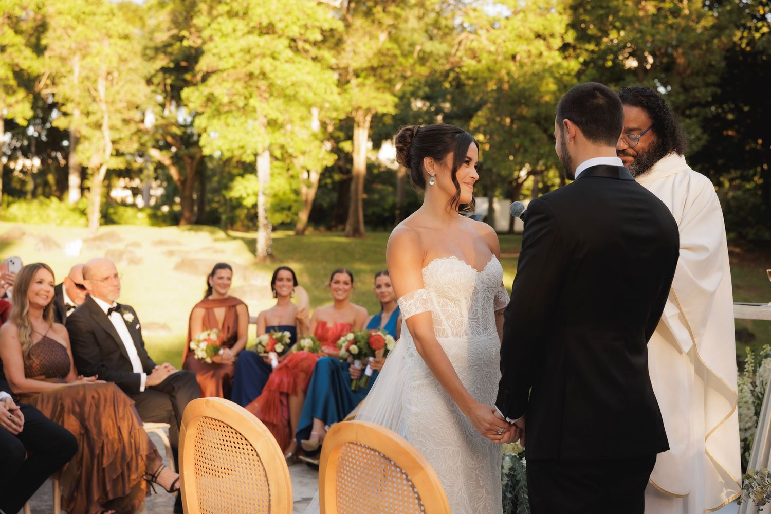 Outdoor wedding ceremony set in a sunlit park with guests seated on chairs watching the couple at the altar.