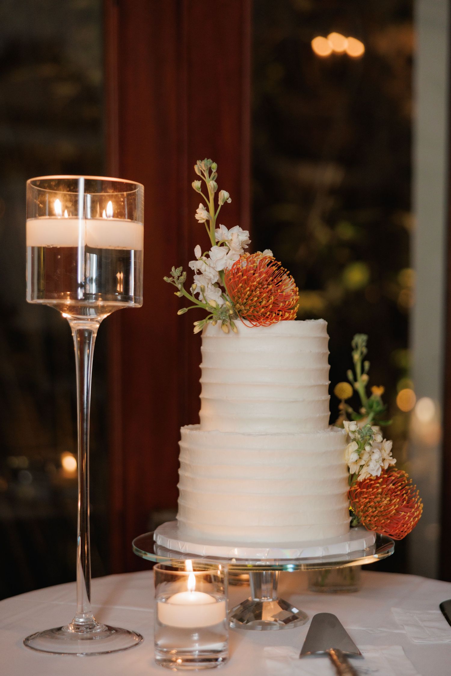 Elegant white two-tier wedding cake decorated with orange protea flowers and delicate white blooms beside tall candleholder.