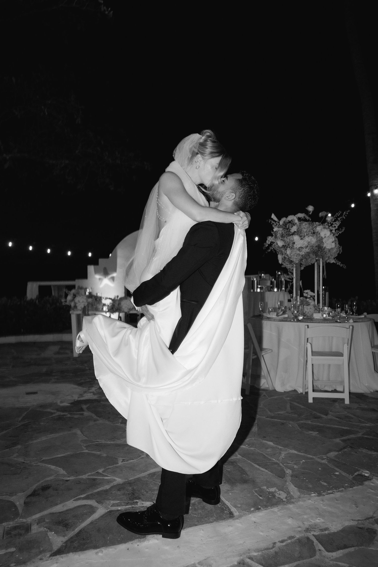 A romantic black and white photograph shows a couple embracing during their first dance at a wedding reception with string lights.