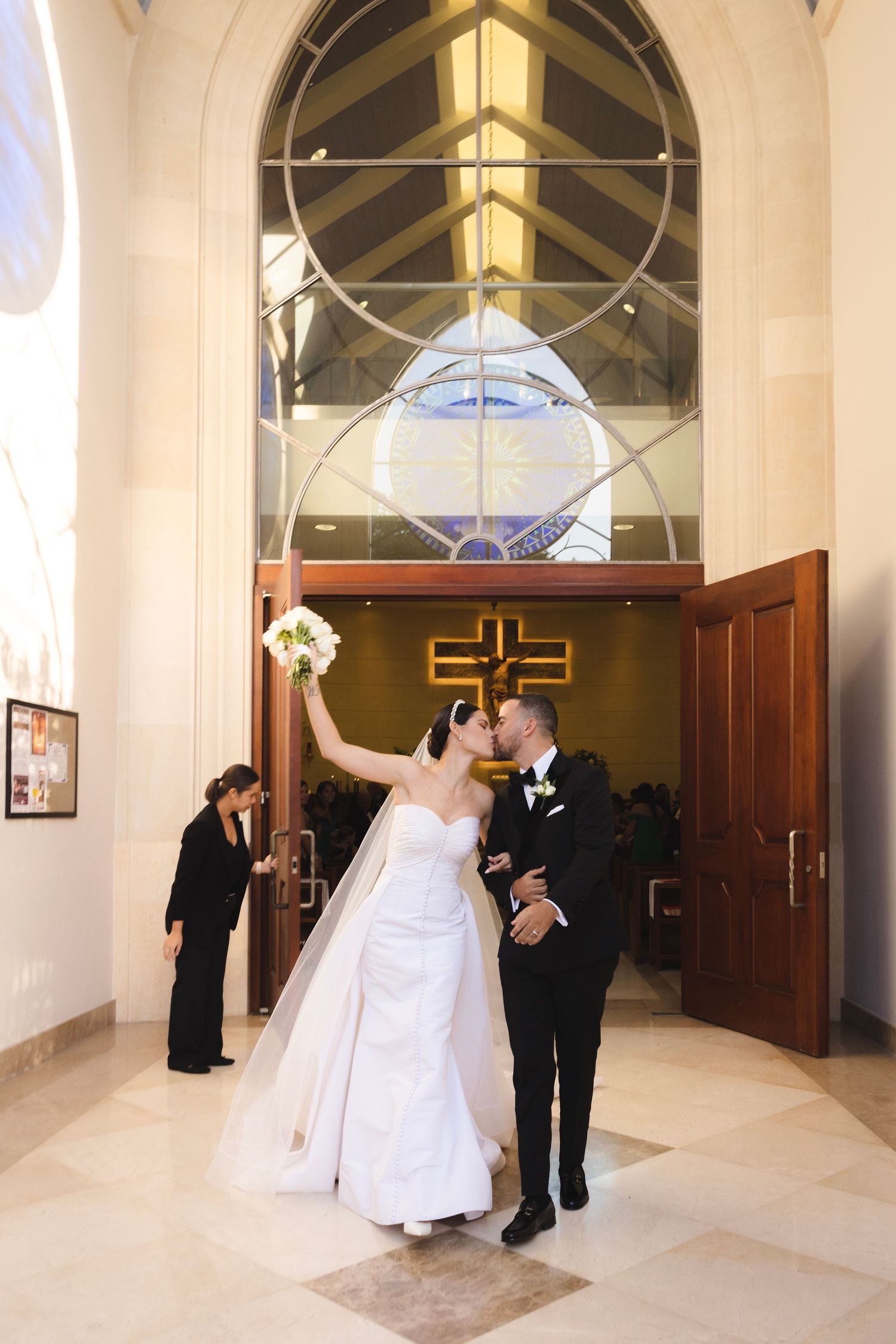 Newlyweds celebrate joyfully while exiting a church chapel with stained glass windows and vaulted ceilings.