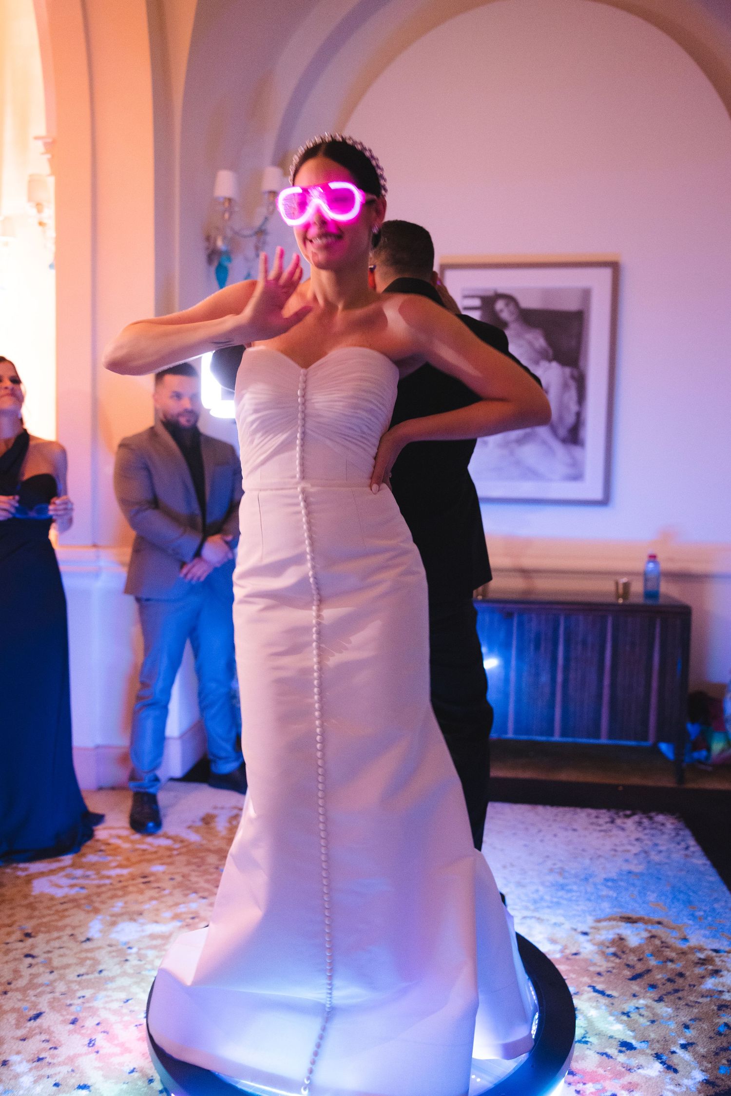 A bride in a white dress and pink heart-shaped glasses strikes a fun pose during her wedding reception.
