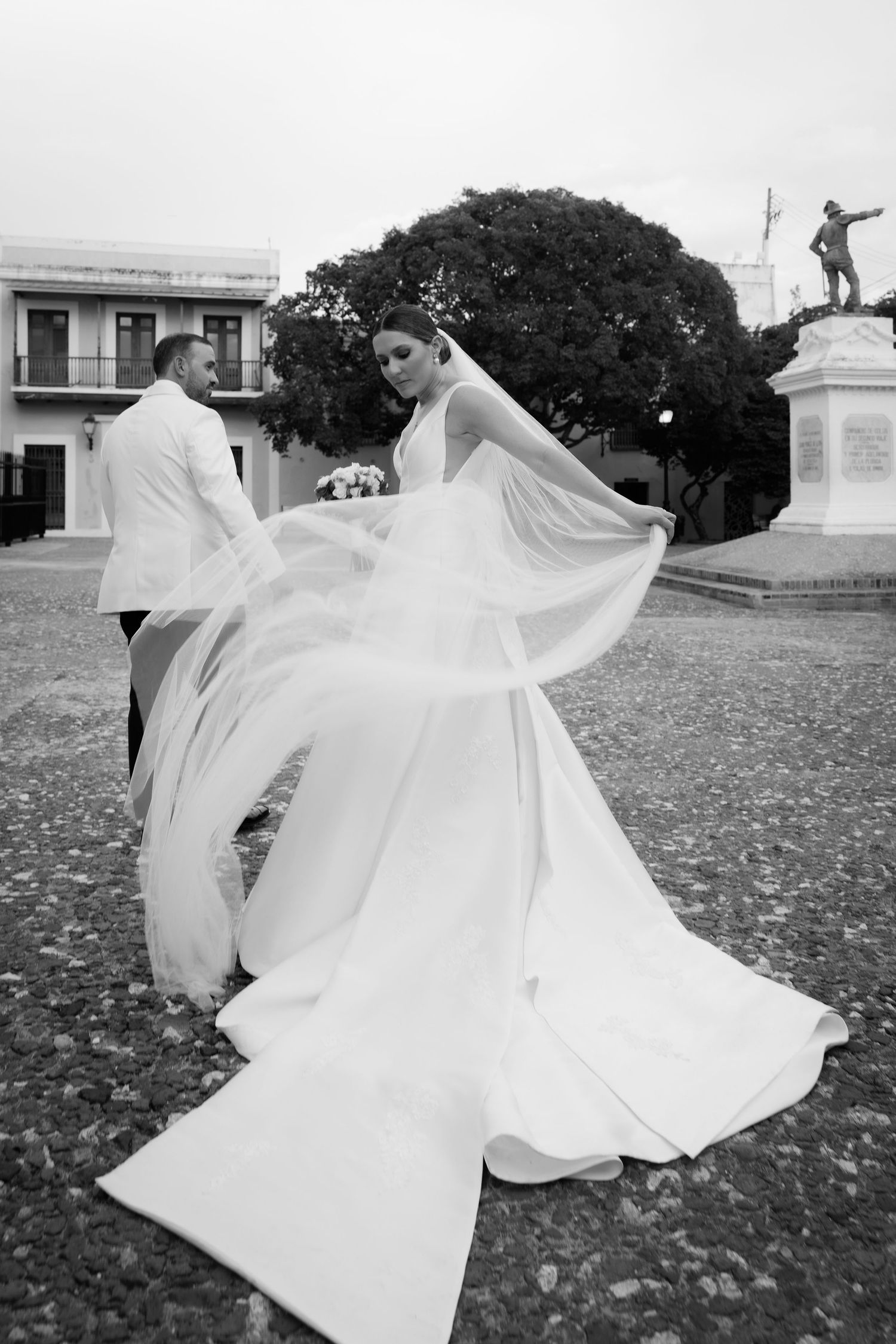 A bride's dress billows dramatically in the wind during an outdoor wedding photoshoot near a statue.
