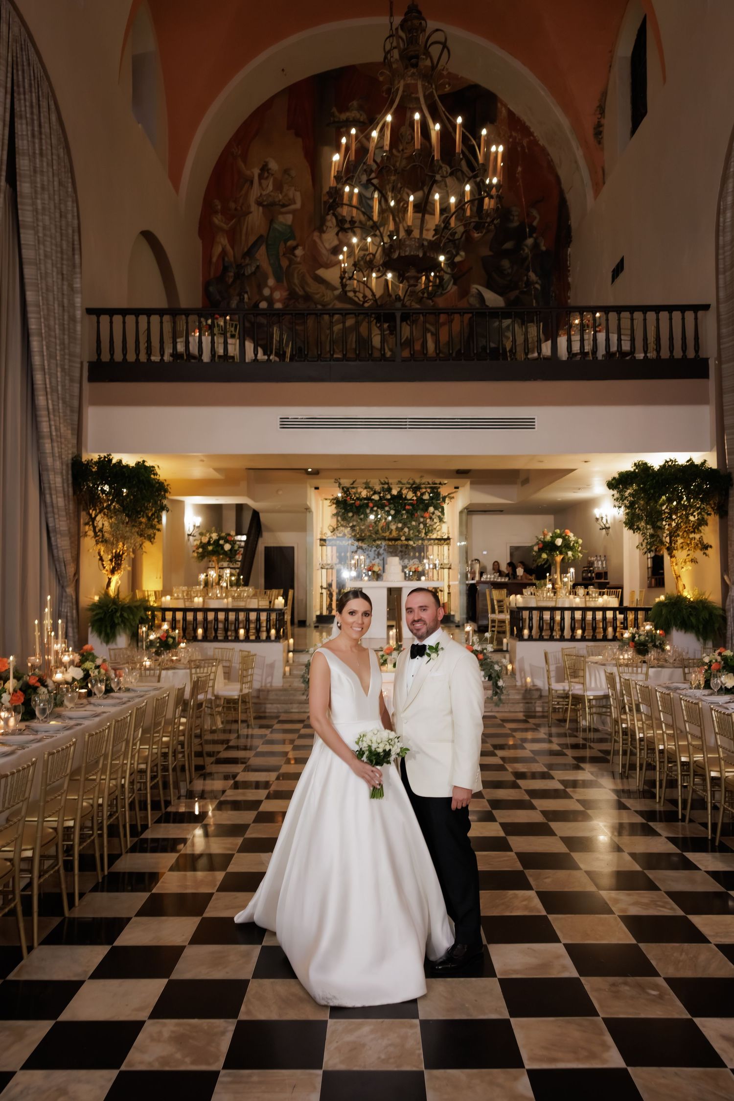Elegant wedding venue with black and white checkered floor, grand chandelier, and ornate balcony in warm lighting.