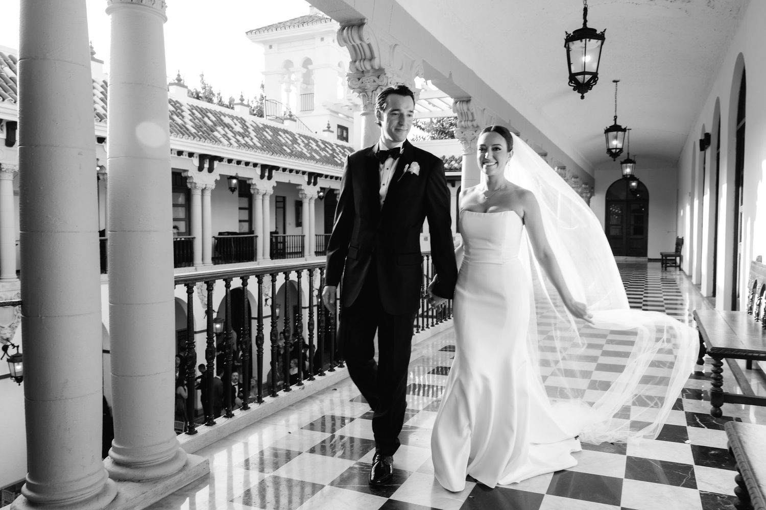 Black and white panoramic wedding photograph showing a couple in formal attire walking along an elegant columned walkway.