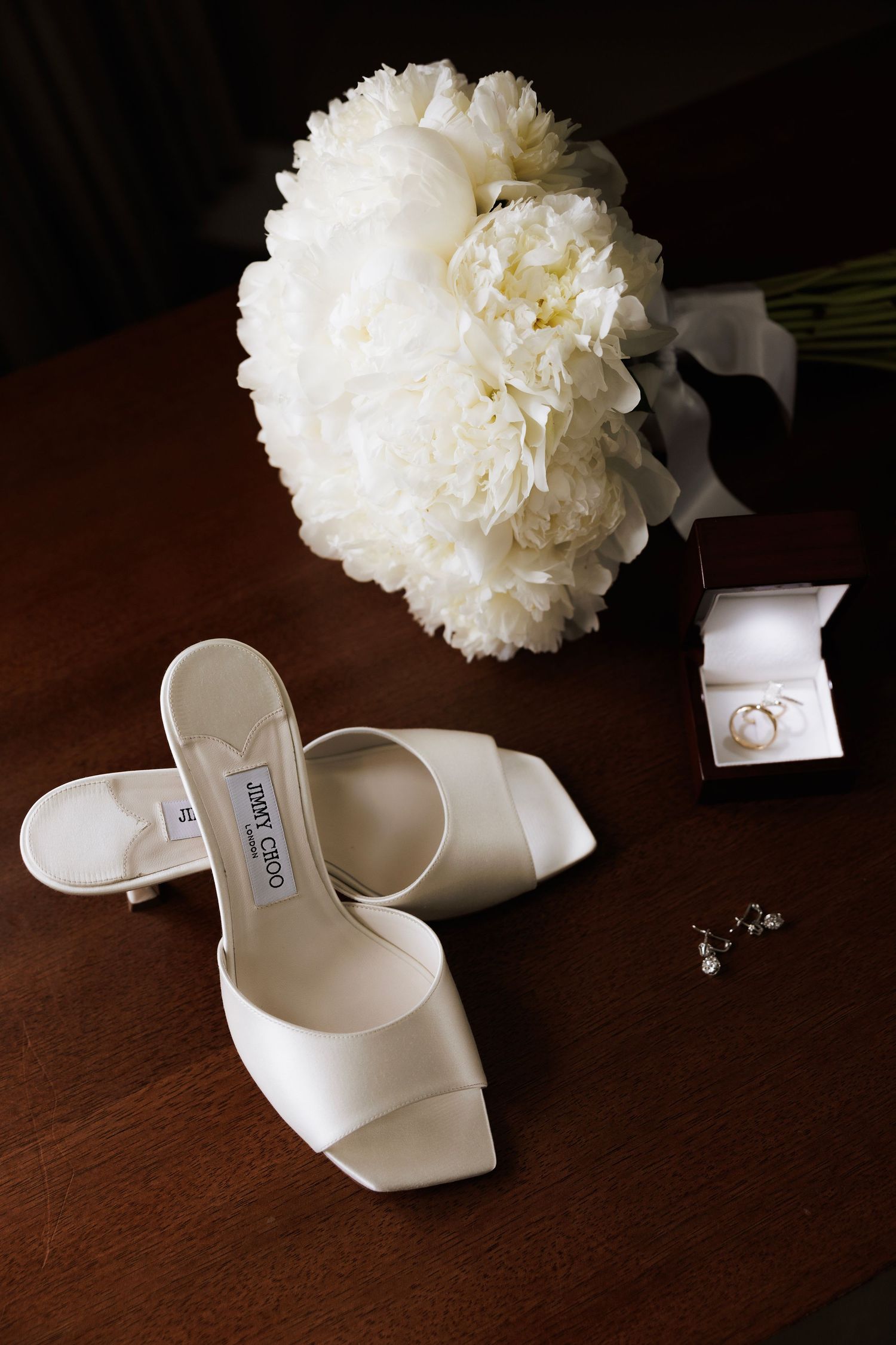 Elegant white bridal shoes paired with a delicate white peony flower on a dark wooden surface.