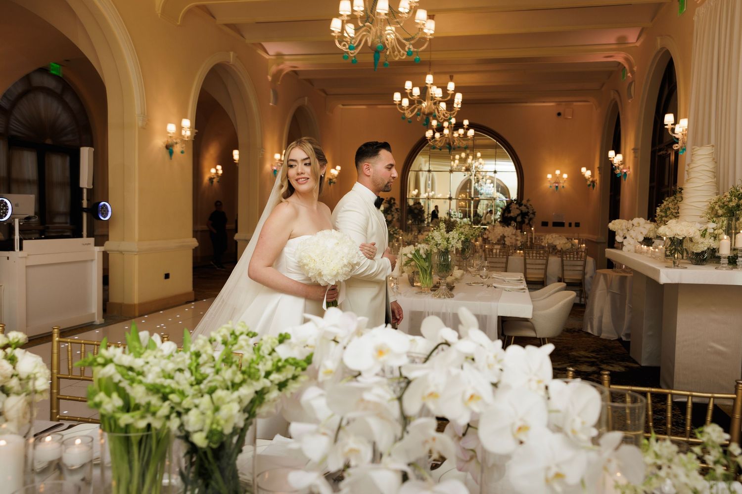 Elegant wedding reception tables decorated with white orchids and crystal chandeliers in a luxurious ballroom setting.