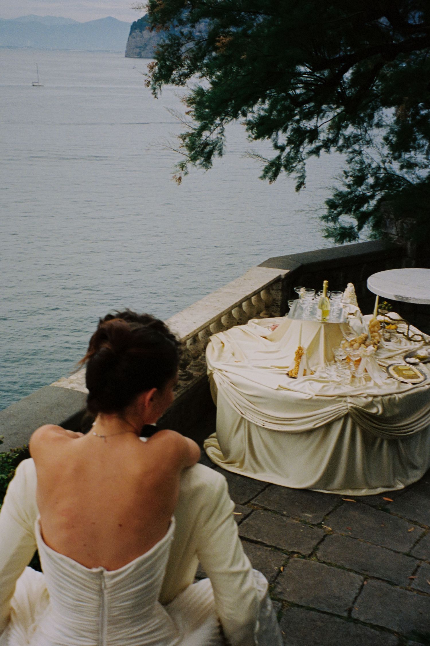 Bride in a white strapless gown sits at an elegantly set table overlooking a serene lake at her wedding.
