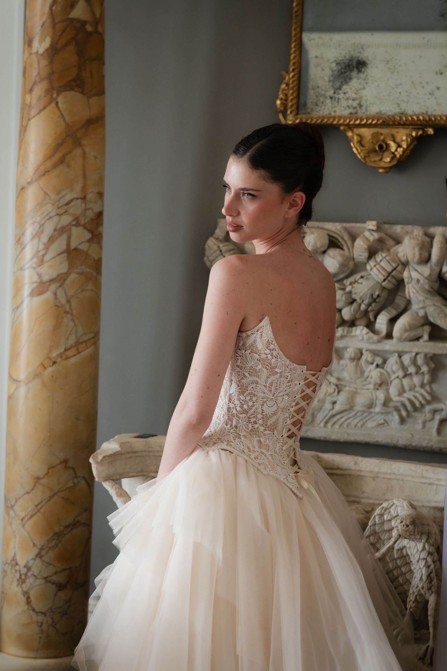 Bride in strapless lace corset ballgown with tulle skirt poses near ornate marble fireplace in elegant interior.