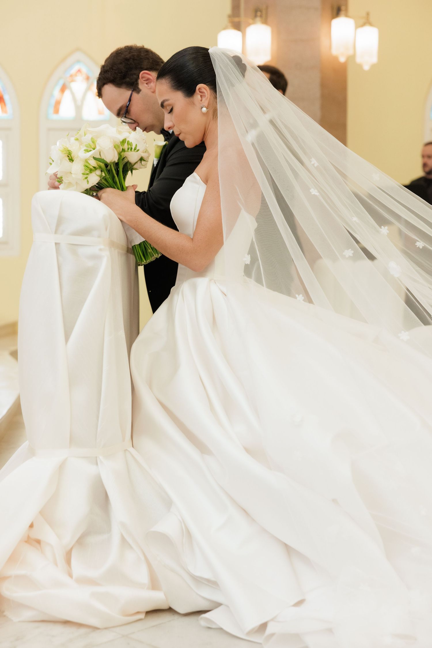 An intimate wedding moment captured in a church with a flowing white dress and dramatic veil.