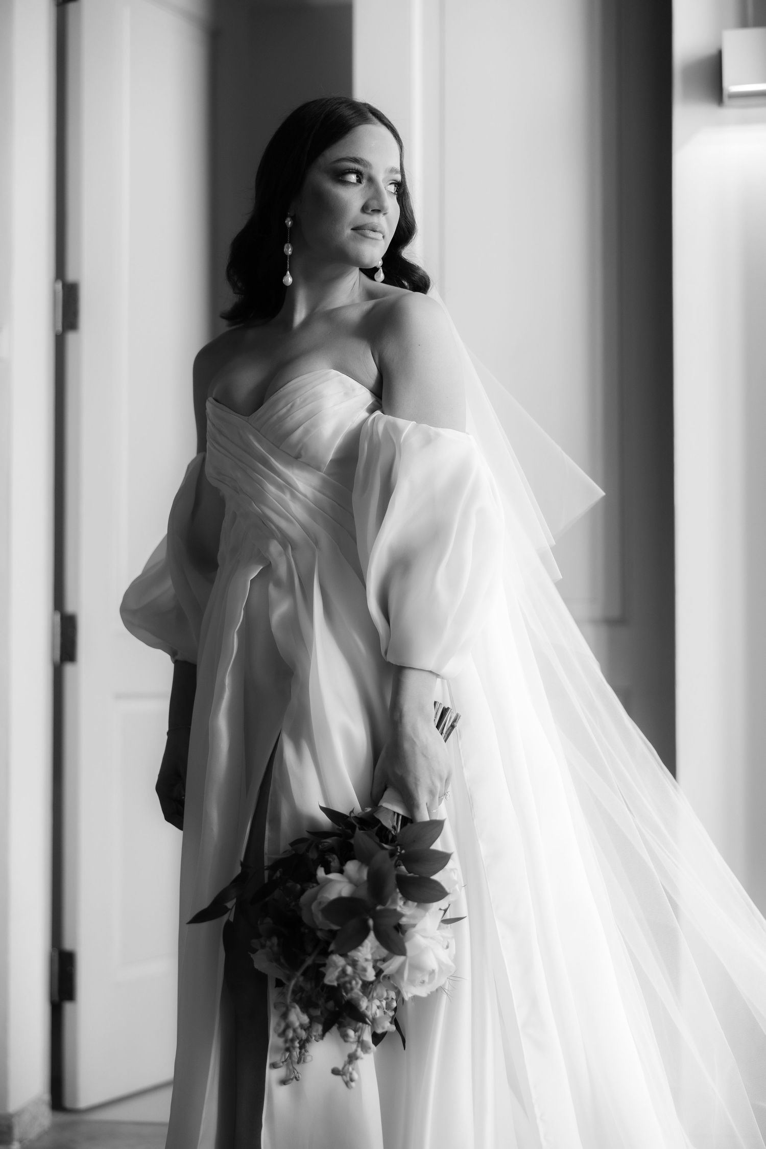 Bride in elegant white dress holding a bouquet, captured in dramatic window light.