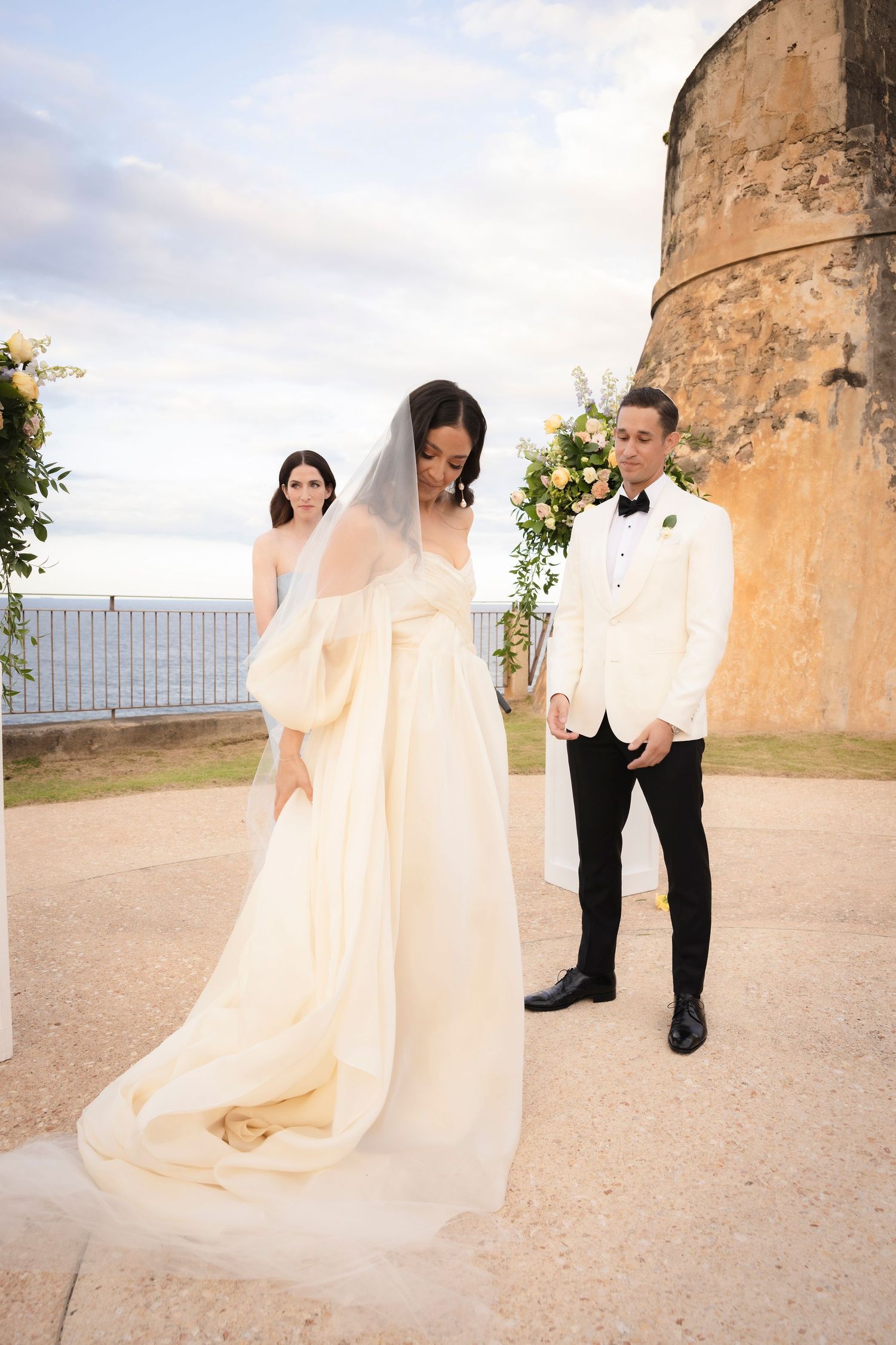 A flowing cream-colored wedding dress billows elegantly against stone pillars in an outdoor setting.
