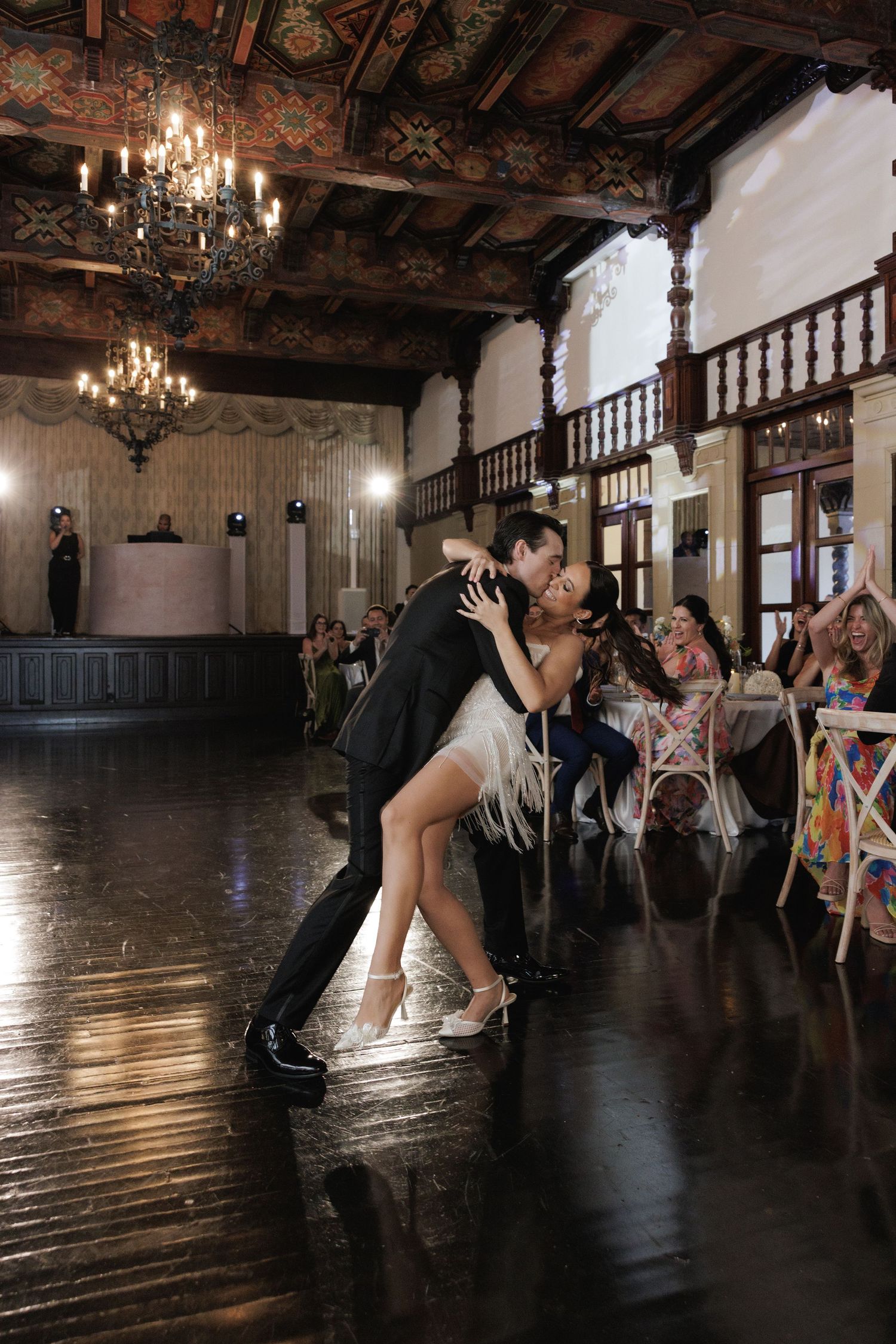 A couple shares a dramatic dance dip on a dark wooden floor in an ornate ballroom with chandeliers.