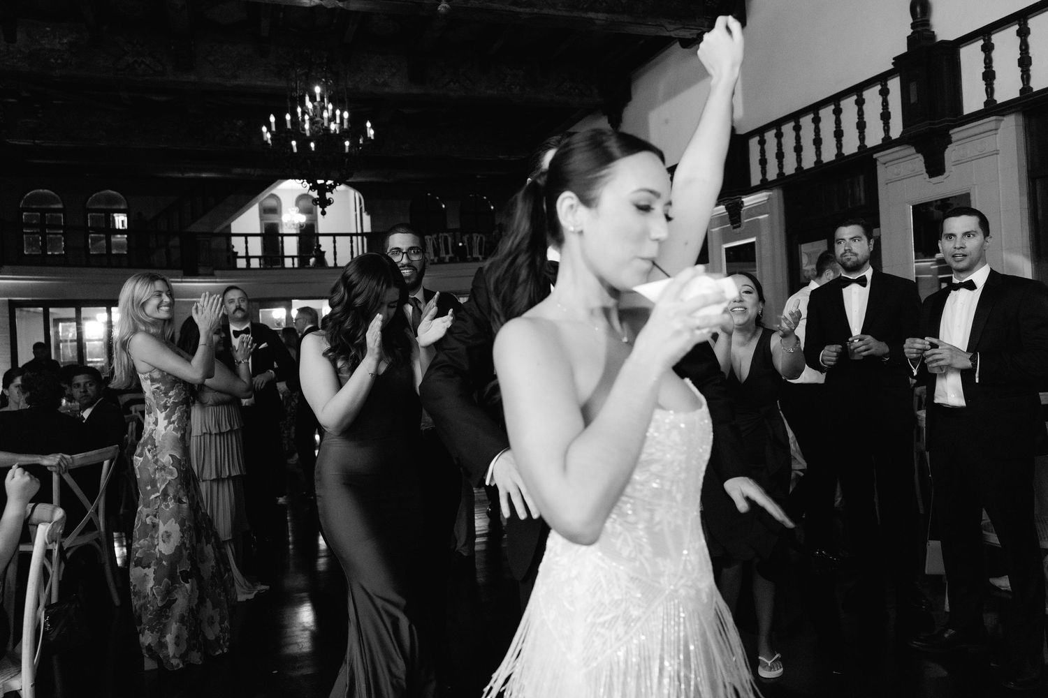 A bride in a white gown dances with one arm raised at an elegant reception venue with guests in formal attire.