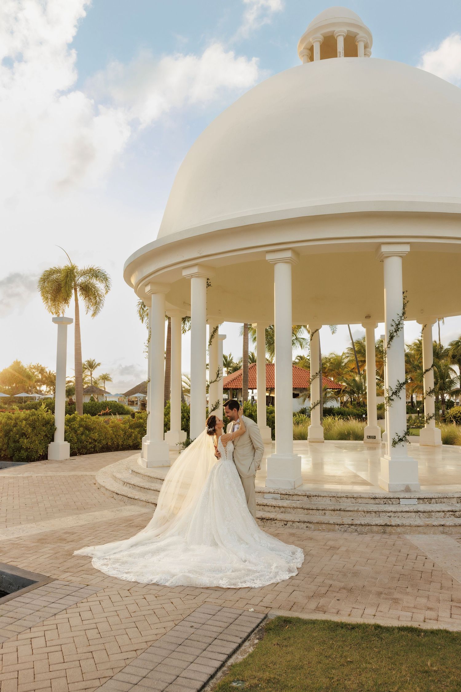 A wedding couple stands in a panoramic view under white columned gazebos at a tropical resort during sunset.