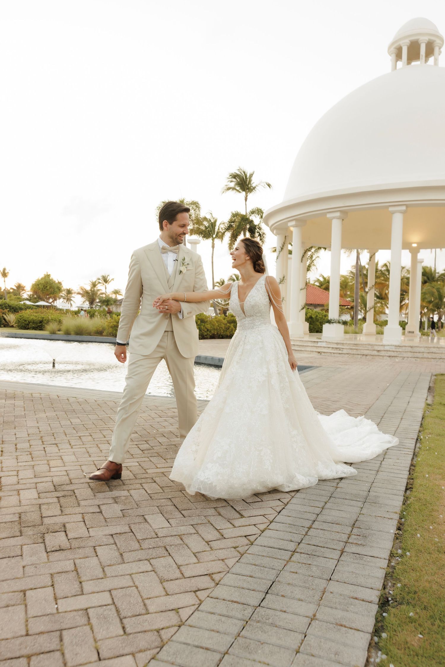 A couple in wedding attire shares a romantic dance on a stone patio near white gazebos and palm trees at sunset.