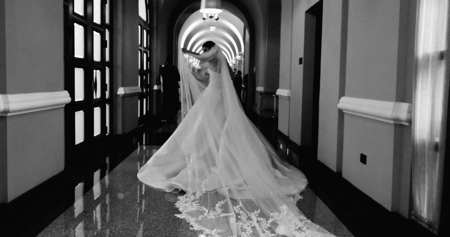 A bride in a flowing white wedding dress appears at a sunlit window in an elegant black and white photograph.