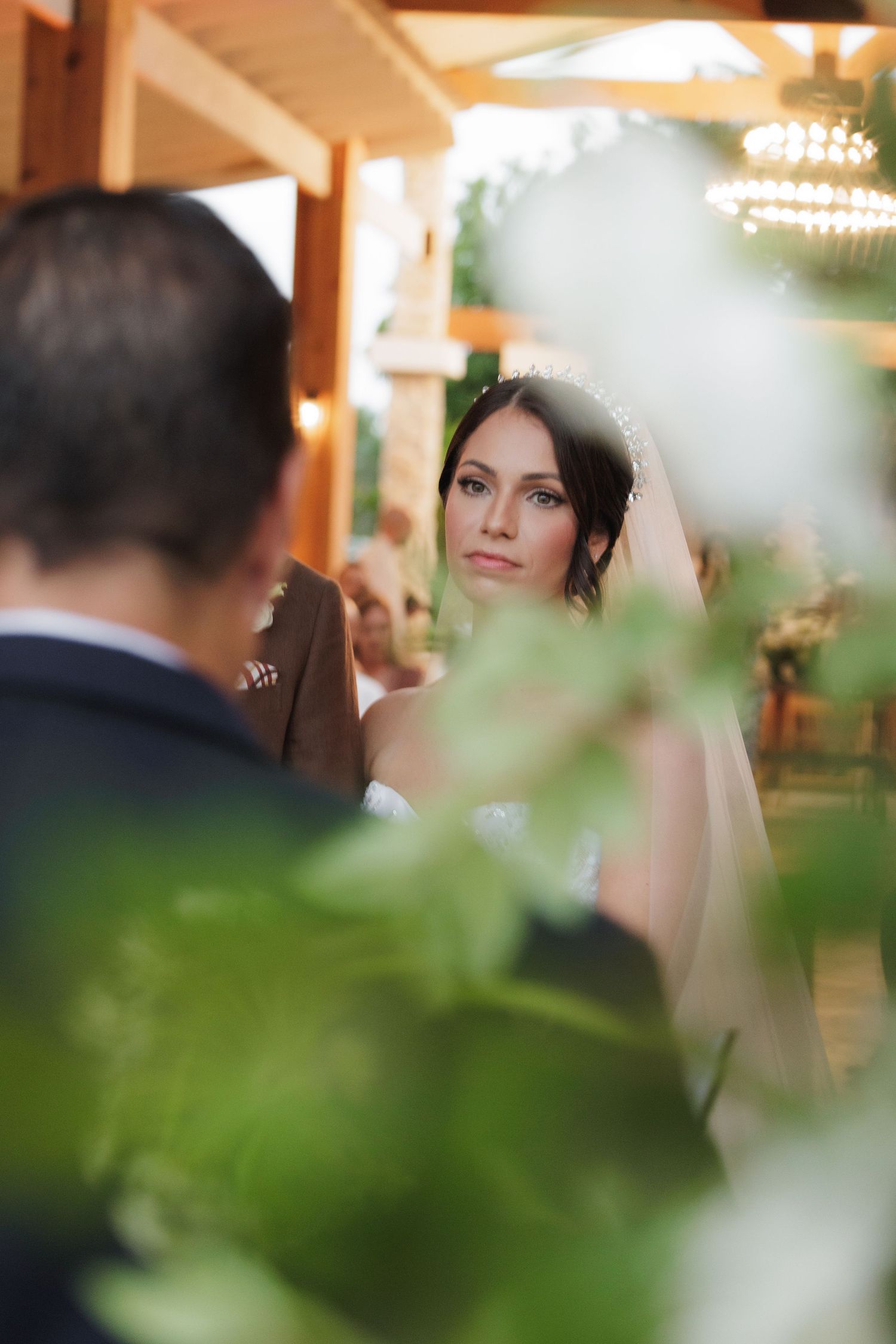 A romantic wedding scene through green foliage with a bride in white veil and elegant decor in warm lighting.