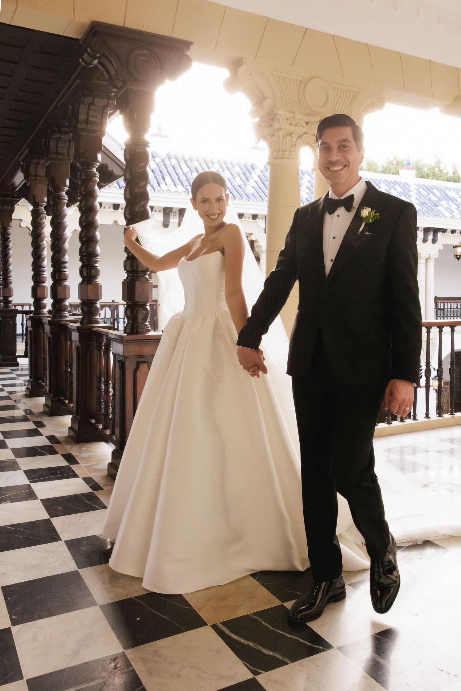 Elegant couple poses on black and white checkered floor under ornate architectural details.