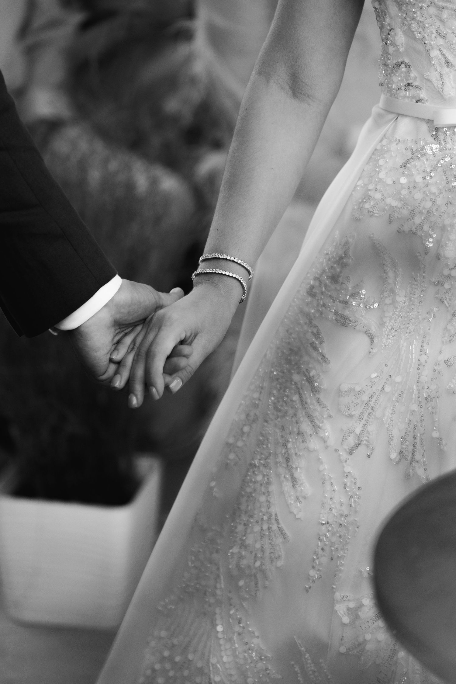 A black and white close-up of newlyweds holding hands, showing the bride's detailed lace dress.