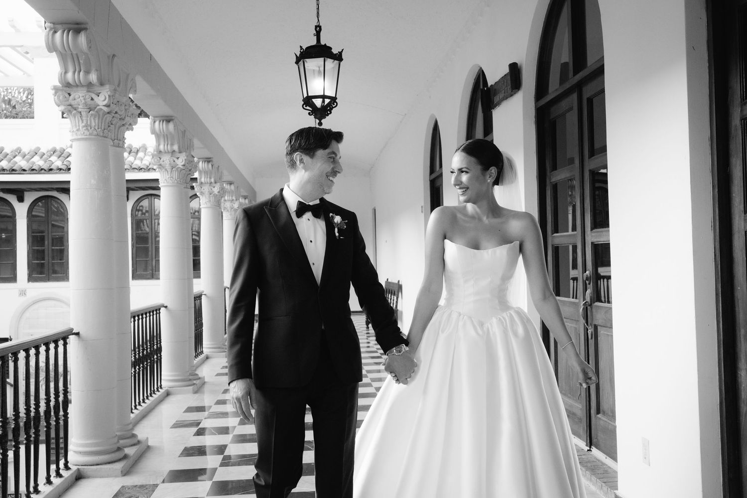 A couple in formal wedding attire share a joyful moment together on a white columned porch.
