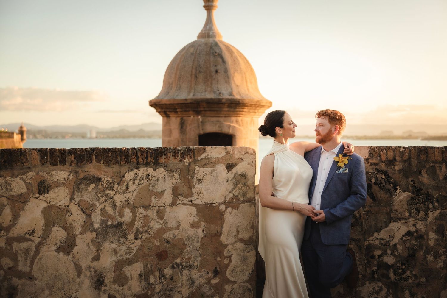 A couple embraces on a stone wall overlooking the ocean during sunset at a historic fortress.