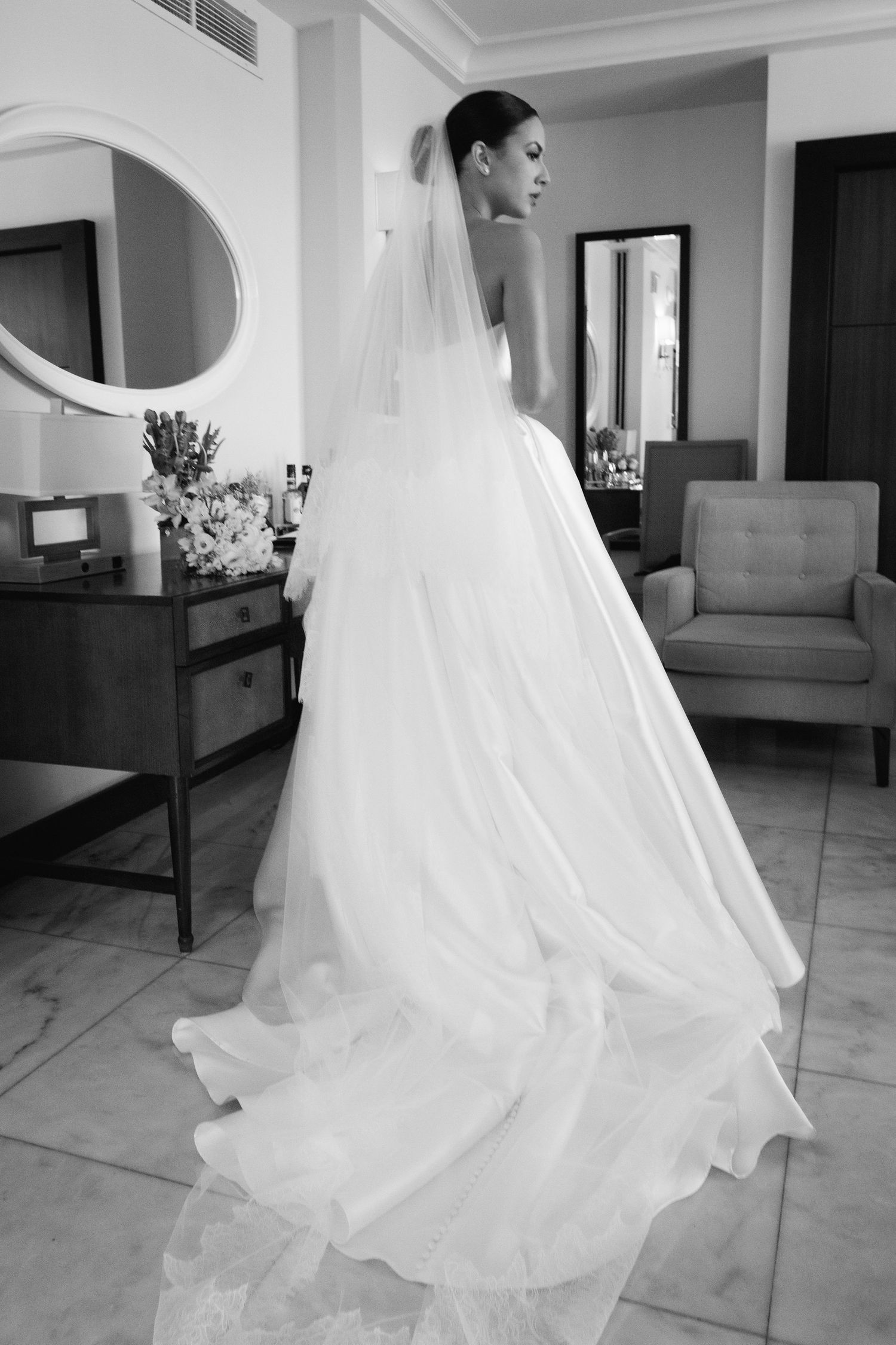 Elegant bride in a flowing white wedding gown and veil poses in a sunlit room with classic decor.
